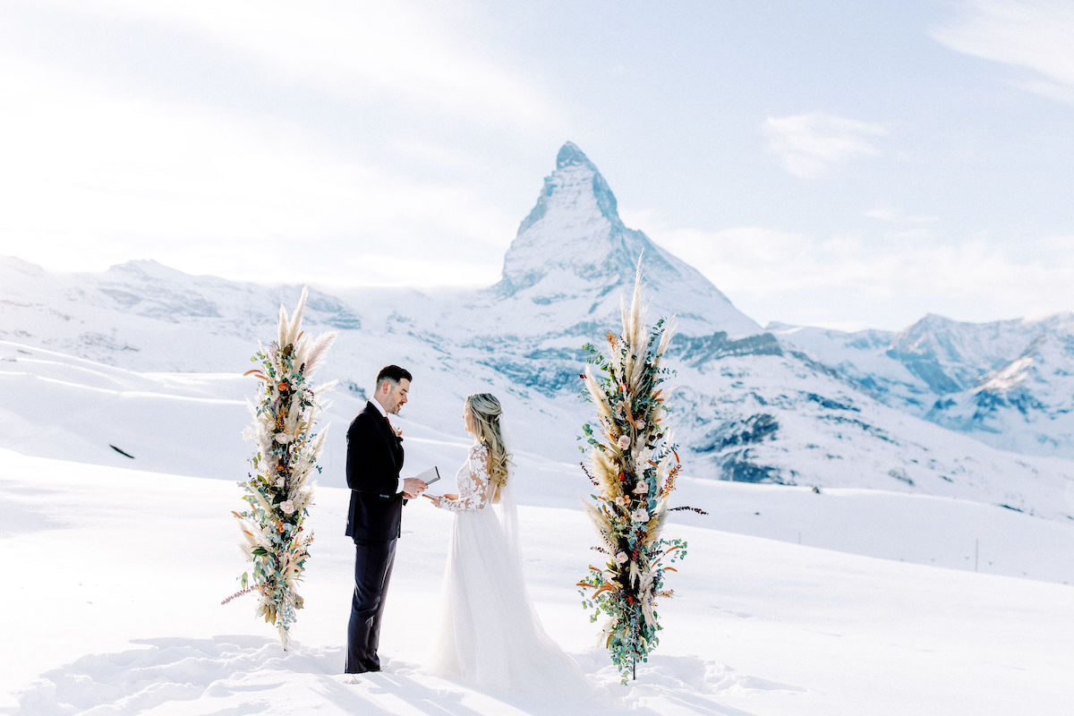 A bride and groom stand facing each other during an outdoor wedding ceremony in a snowy mountain landscape with floral arrangements and a peak in the background.