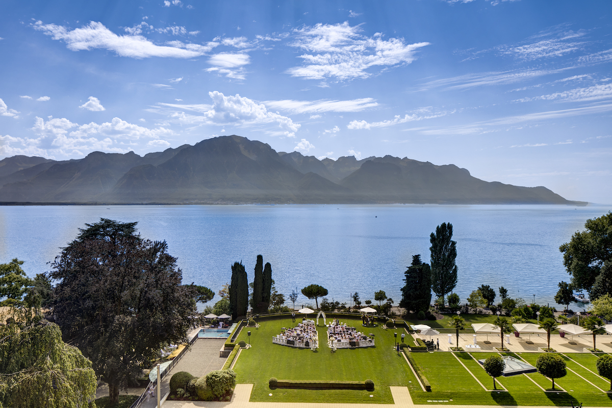 A landscaped garden with paths and trees borders a calm lake, with mountains visible in the background under a partly cloudy sky.