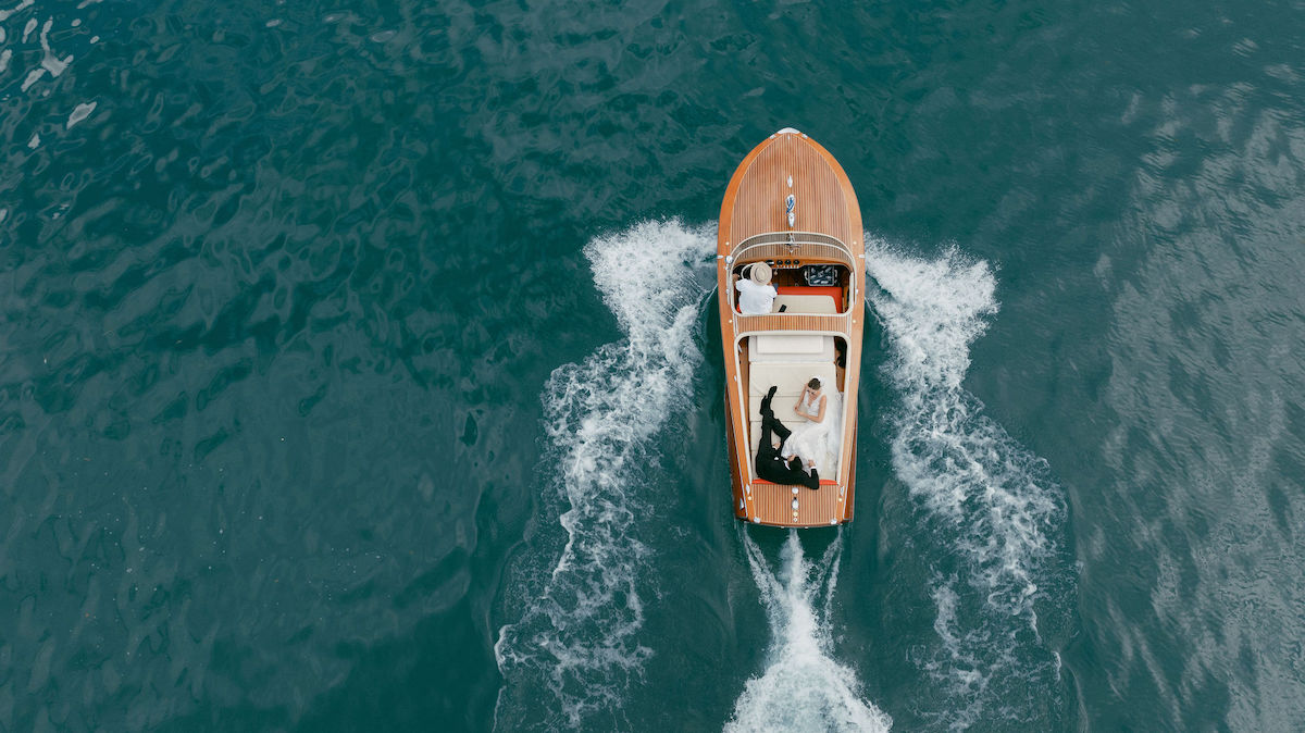 Aerial view of a wooden speedboat moving through blue water with a couple dressed in formal attire lying on the deck.