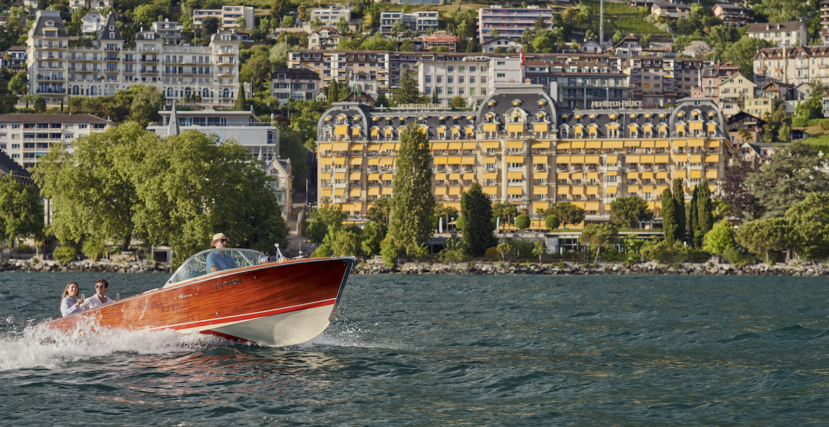 A red wooden speedboat with three people travels on a lake in front of a large yellow hotel and hillside buildings.