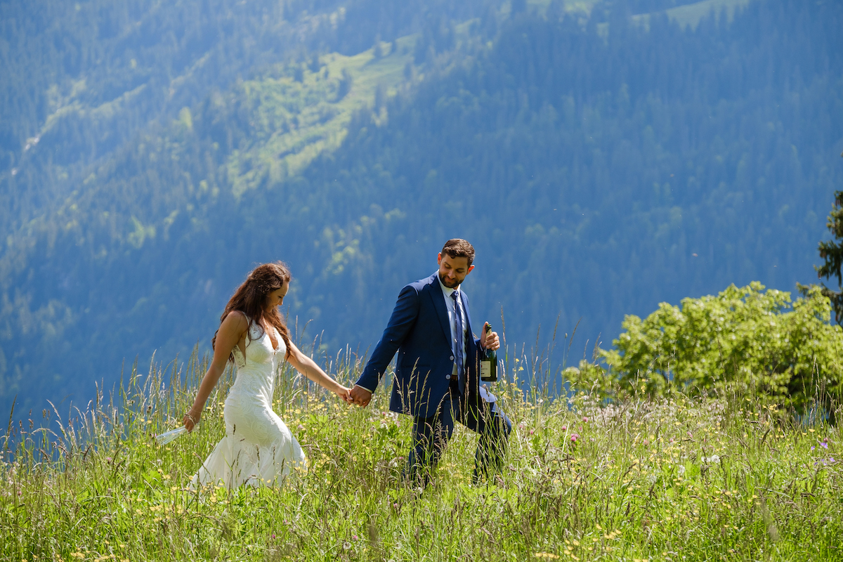 A couple in formal attire walks hand in hand through tall grass on a hillside with mountains in the background. The man carries a wine bottle.