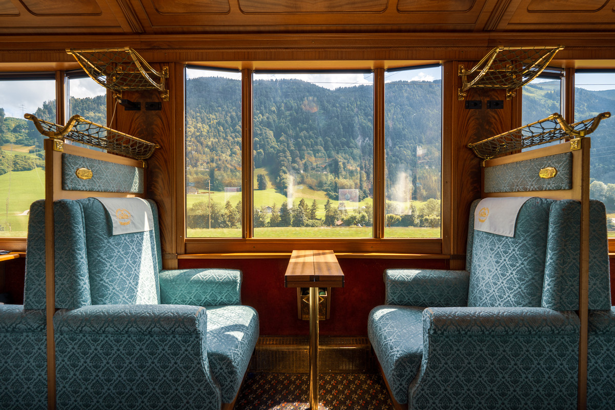 Four upholstered train seats surround a small wooden table inside a vintage railcar, with large windows showing a scenic mountain and countryside view.