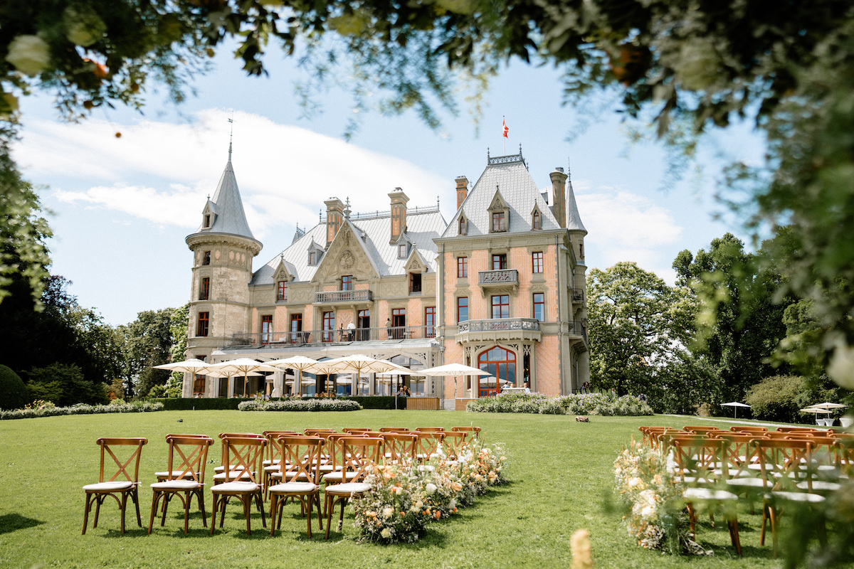 Rows of wooden chairs and floral arrangements are set up on a lawn facing a large, ornate castle-like building under a partly cloudy sky.