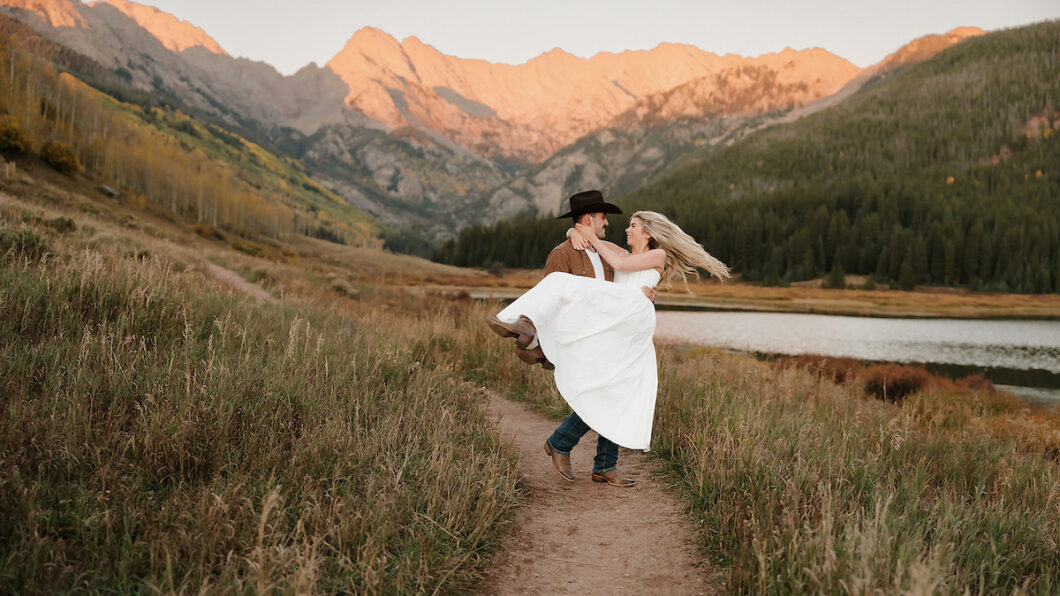A man in a cowboy hat lifts a woman in a white dress on a grassy path with mountains and a lake in the background at sunset.