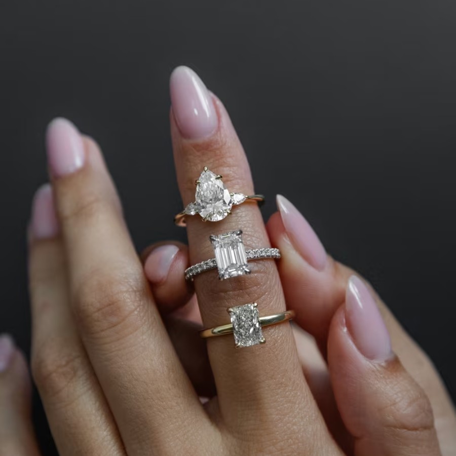 A hand with manicured nails displays three diamond rings of different shapes and bands on the index, middle, and ring fingers against a dark background.