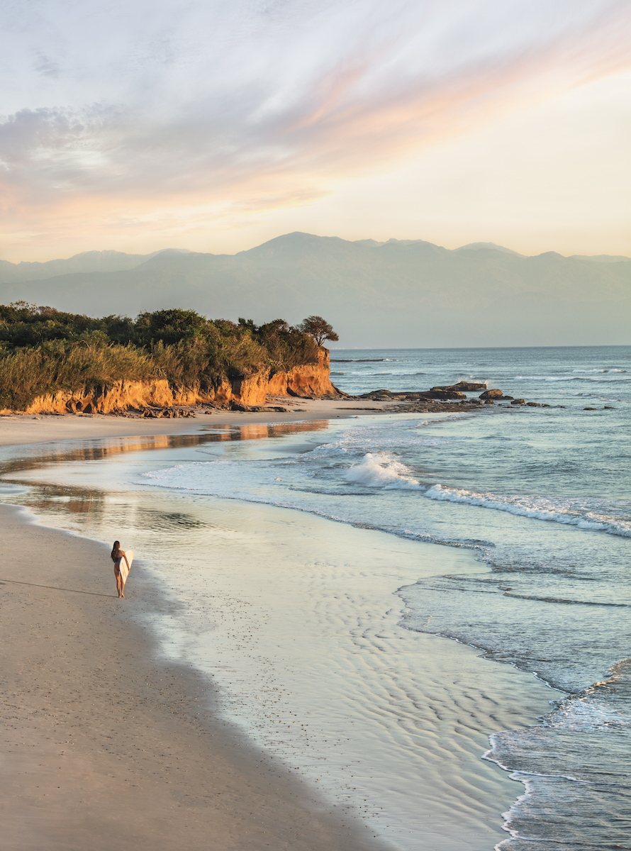 A person walks alone along a sandy beach at sunset, with waves gently rolling in and cliffs and trees in the background.