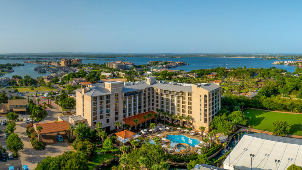 Aerial view of a large hotel with multiple floors, a courtyard swimming pool, palm trees, and a lake with surrounding buildings in the background.