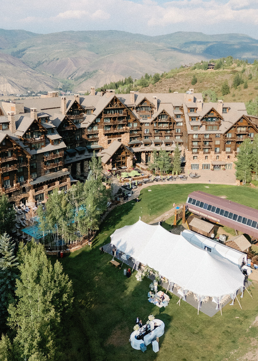 A large rustic lodge surrounded by mountains and trees, with a white event tent set up on a grassy lawn for an outdoor gathering.