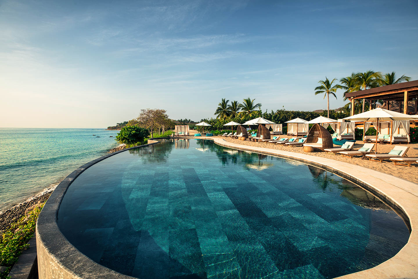 Infinity pool overlooking the ocean with lounge chairs and umbrellas along the poolside, surrounded by palm trees under a clear sky.