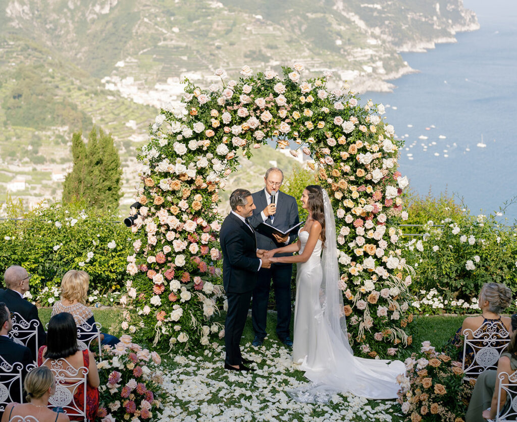 A couple stands under a large floral arch exchanging vows during an outdoor wedding ceremony overlooking a coastal landscape.
