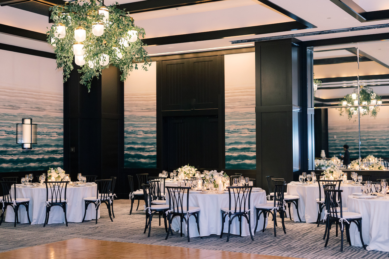 Elegant banquet hall with round tables set for an event, featuring white tablecloths, floral centerpieces, and black chairs, with chandeliers and wall art depicting ocean waves.