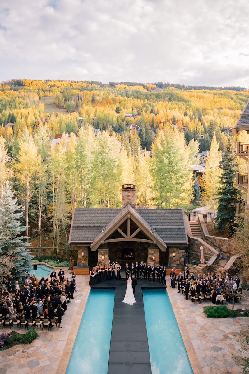 A wedding ceremony takes place outdoors by a rectangular pool, surrounded by guests and trees with autumn foliage in a mountainous area.