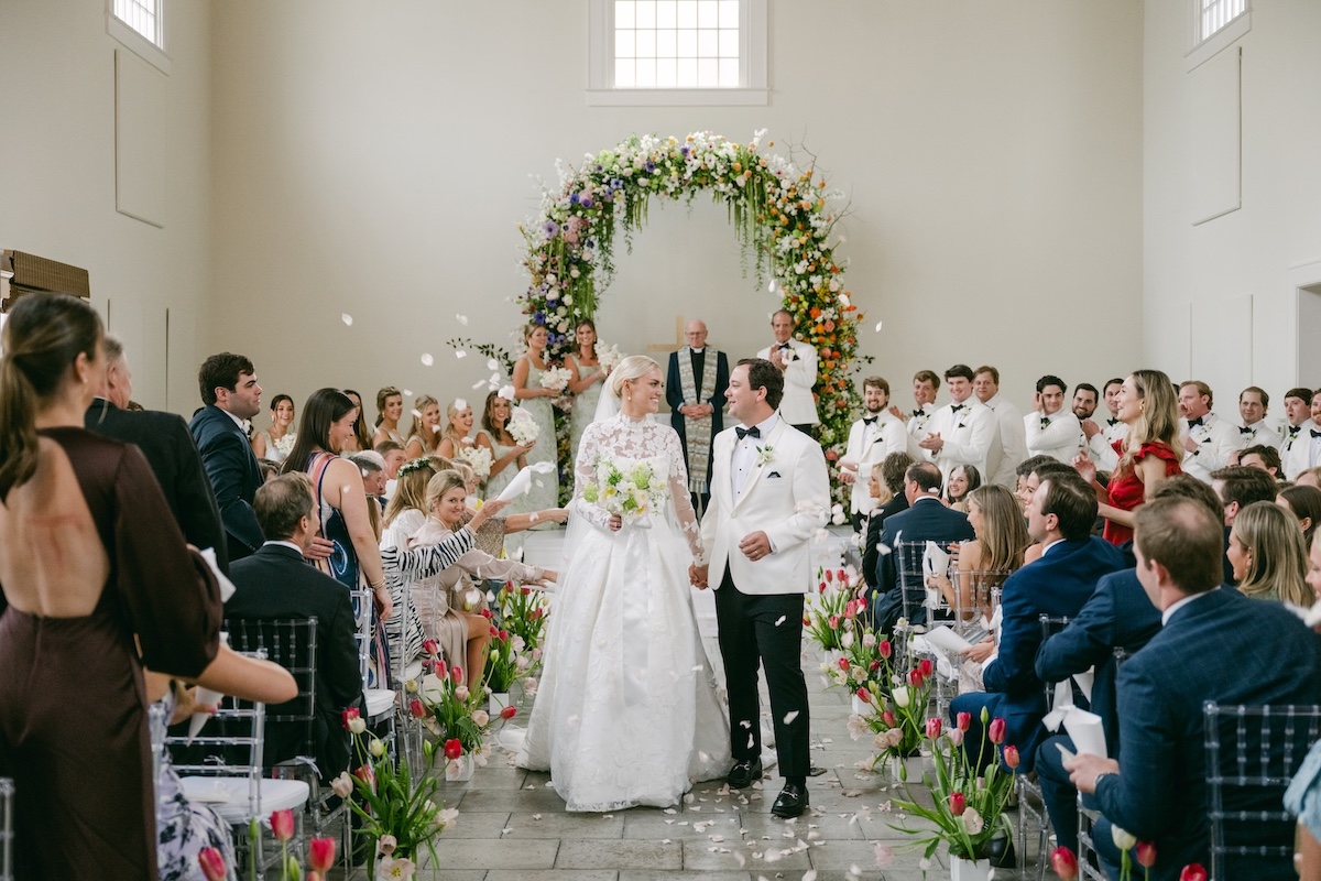 A bride and groom walk down the aisle, smiling, as guests throw flower petals in a decorated indoor wedding ceremony with floral arrangements and an arch.