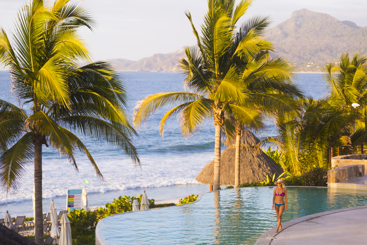 A woman in a swimsuit stands at the edge of an infinity pool overlooking the ocean, with palm trees and mountains in the background.