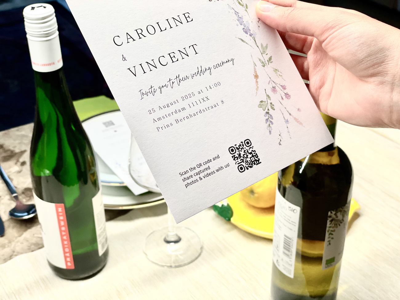 A hand holds a wedding invitation card above a table with two wine bottles, a wine glass, and plates.
