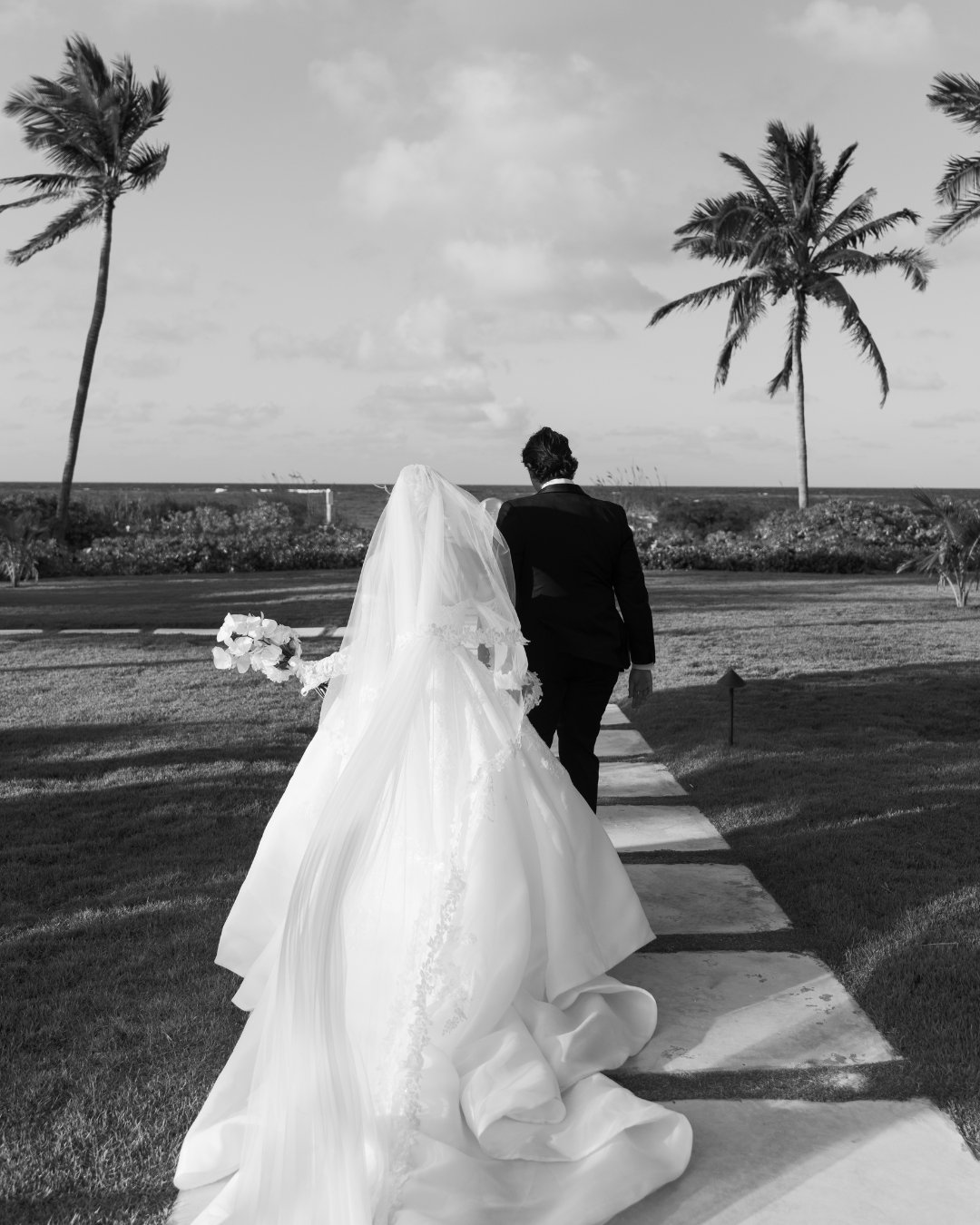 A bride and groom walk down a paved path outdoors, with palm trees and the ocean in the background. The photo is in black and white.