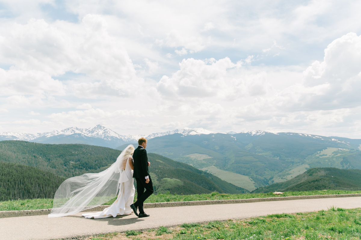A bride and groom walk outside on a paved path with mountains and forests in the background under a partly cloudy sky.