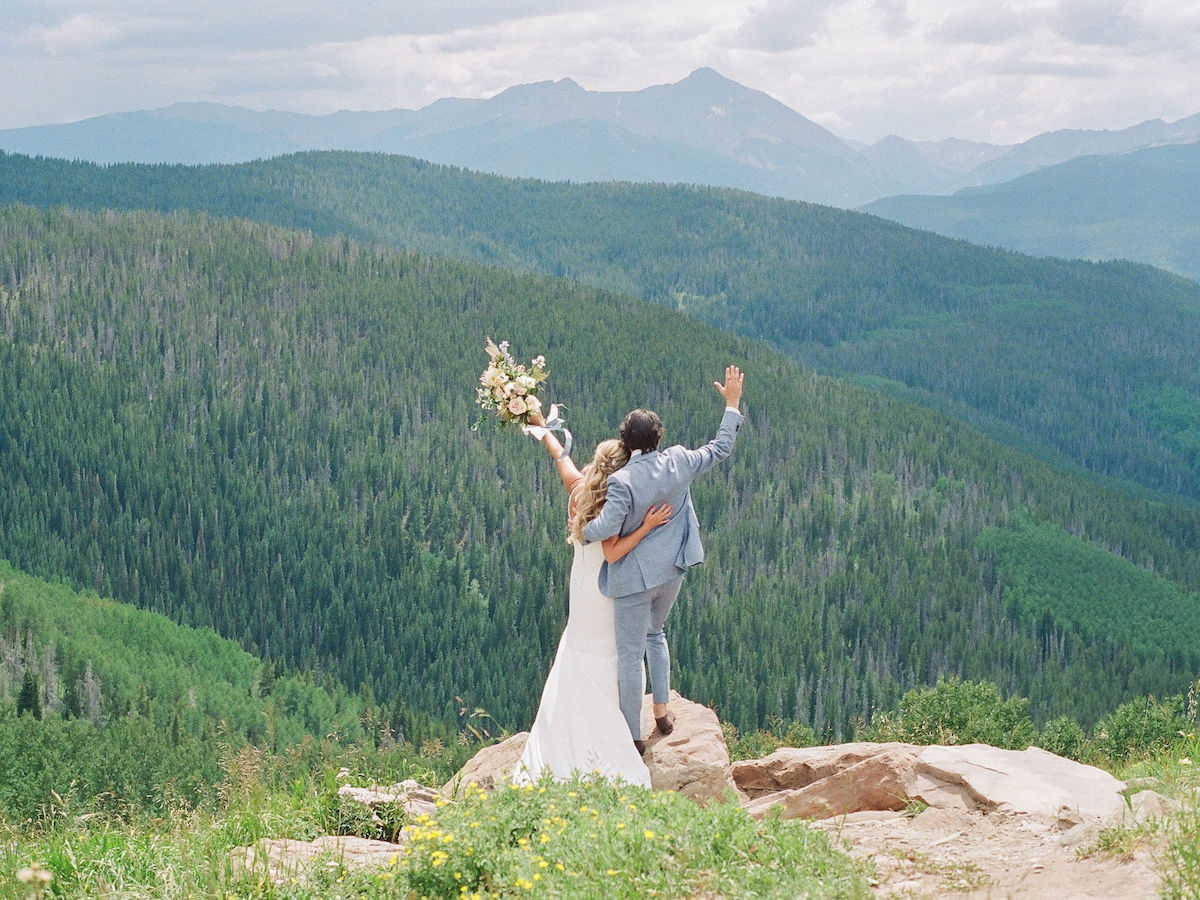 A couple in wedding attire stands on a rock, facing a vast green mountain landscape; the woman holds a bouquet and both have an arm raised.