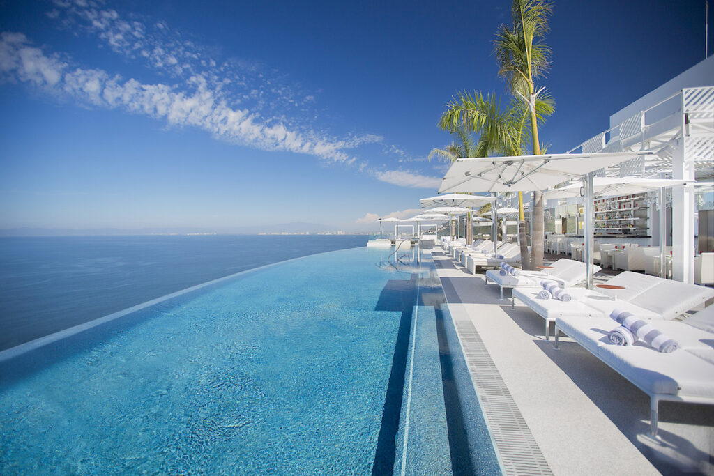 Infinity pool with clear blue water overlooks the ocean, lined with white lounge chairs, umbrellas, and palm trees under a clear sky.