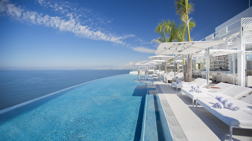 Infinity pool with clear blue water overlooks the ocean, lined with white lounge chairs, umbrellas, and palm trees under a clear sky.