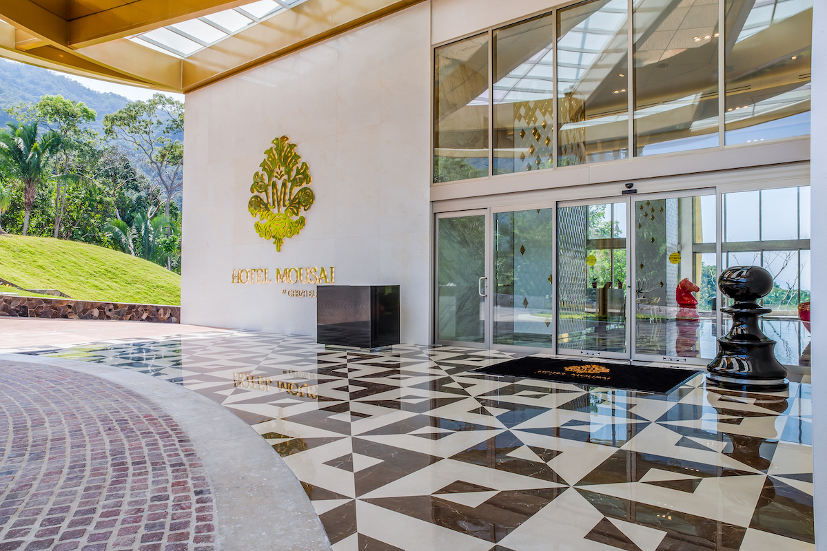 Hotel entrance with large glass doors, geometric black and white tile floor, and gold hotel logo on a white wall; greenery visible outside.