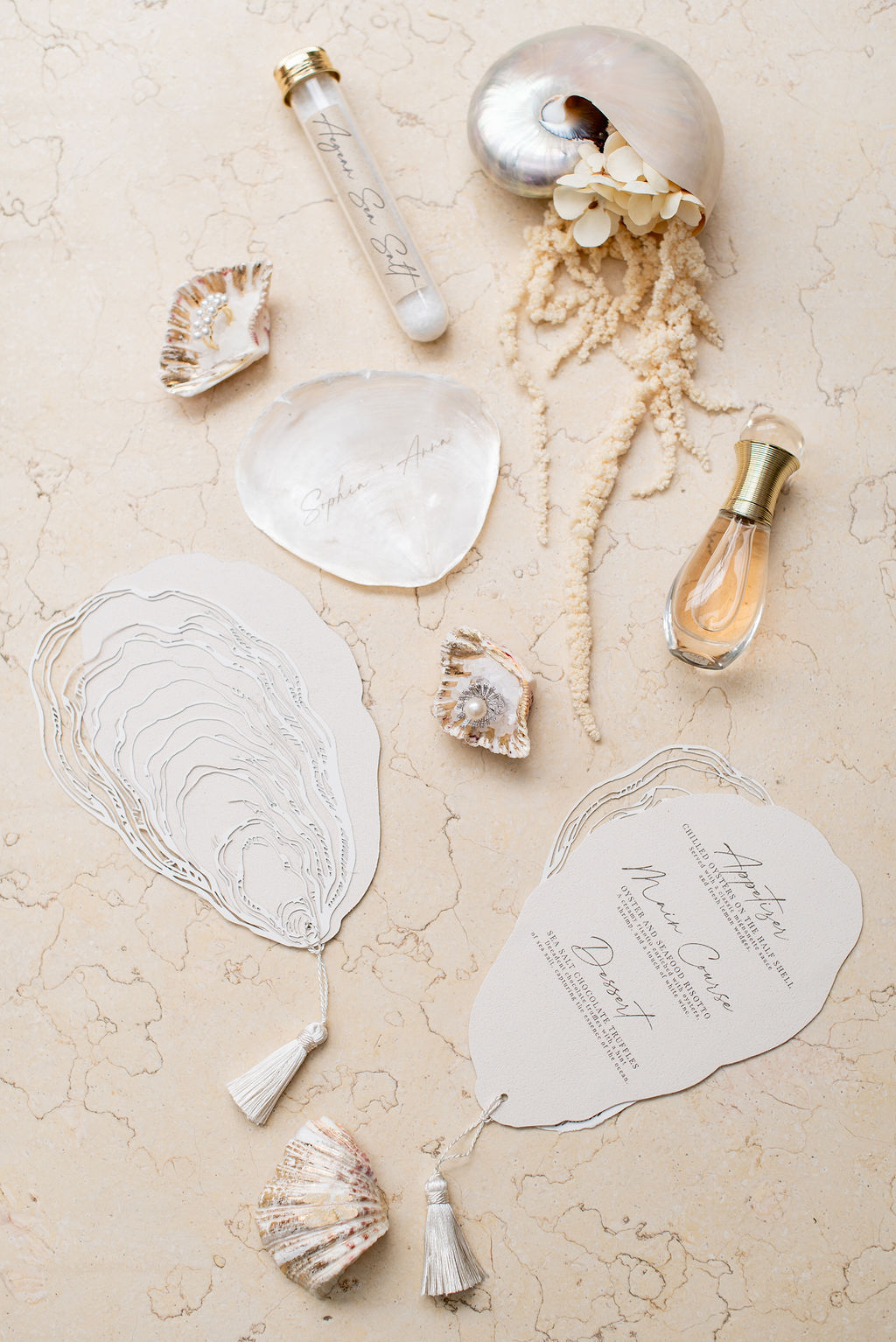 Flat lay of oyster-shaped wedding invitations with tassels, seashells, a pearlized shell, a glass vial, and a small perfume bottle on a beige marble surface.