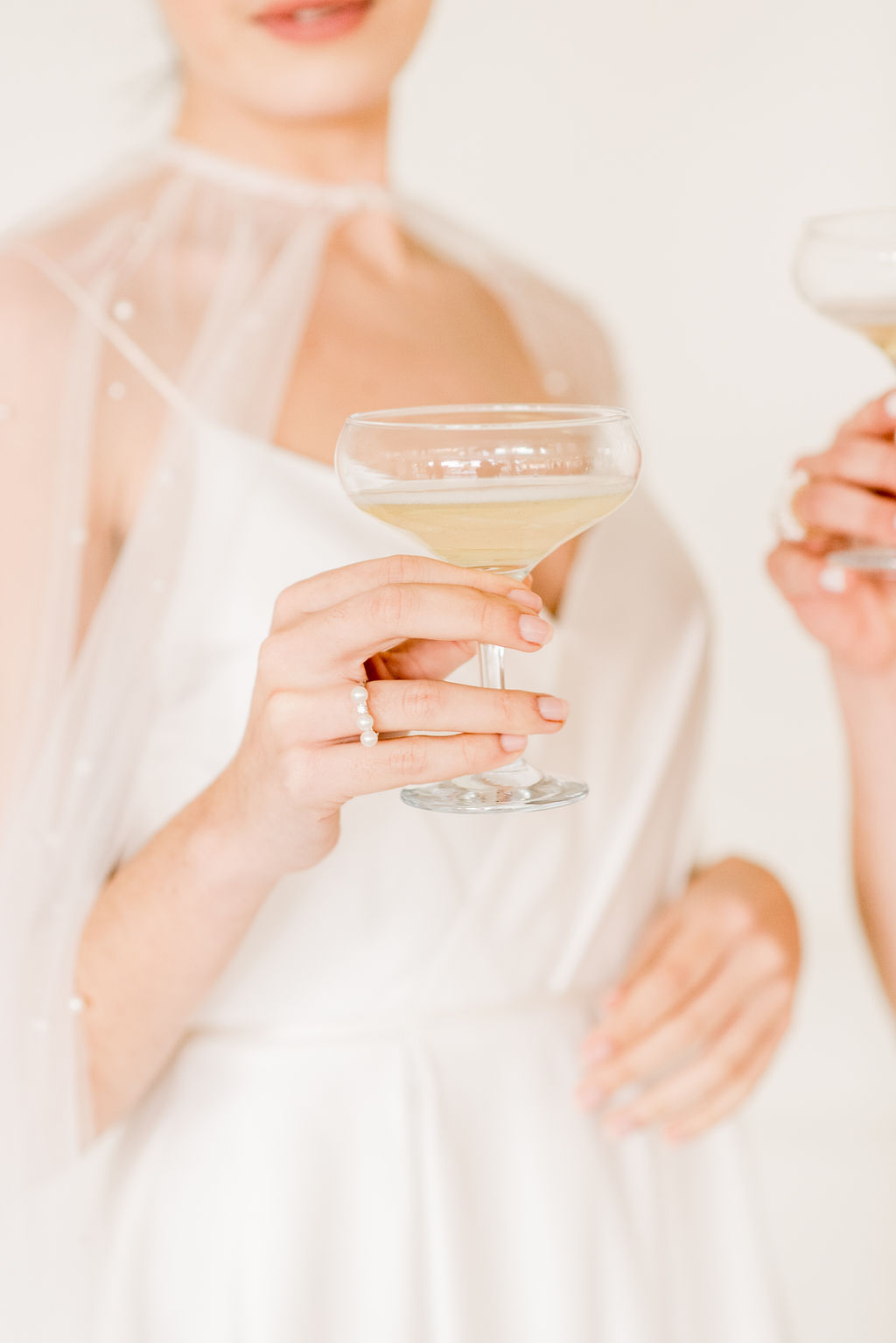 A woman in a white dress holds a coupe glass filled with champagne, with a pearl-detailed sheer cape and an engagement ring visible on her hand. Another person holds a similar glass in the background.