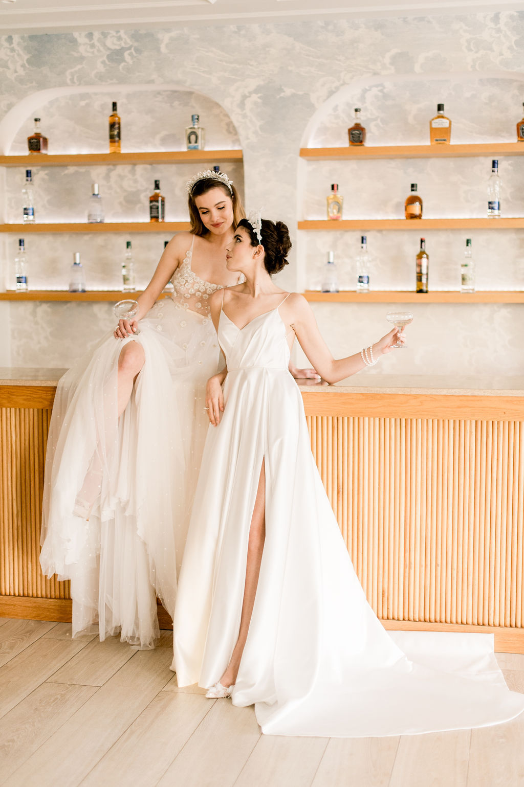 Two women in wedding dresses pose together at a modern bar with bottles on shelves behind them; one sits on the counter while the other stands, holding a glass.
