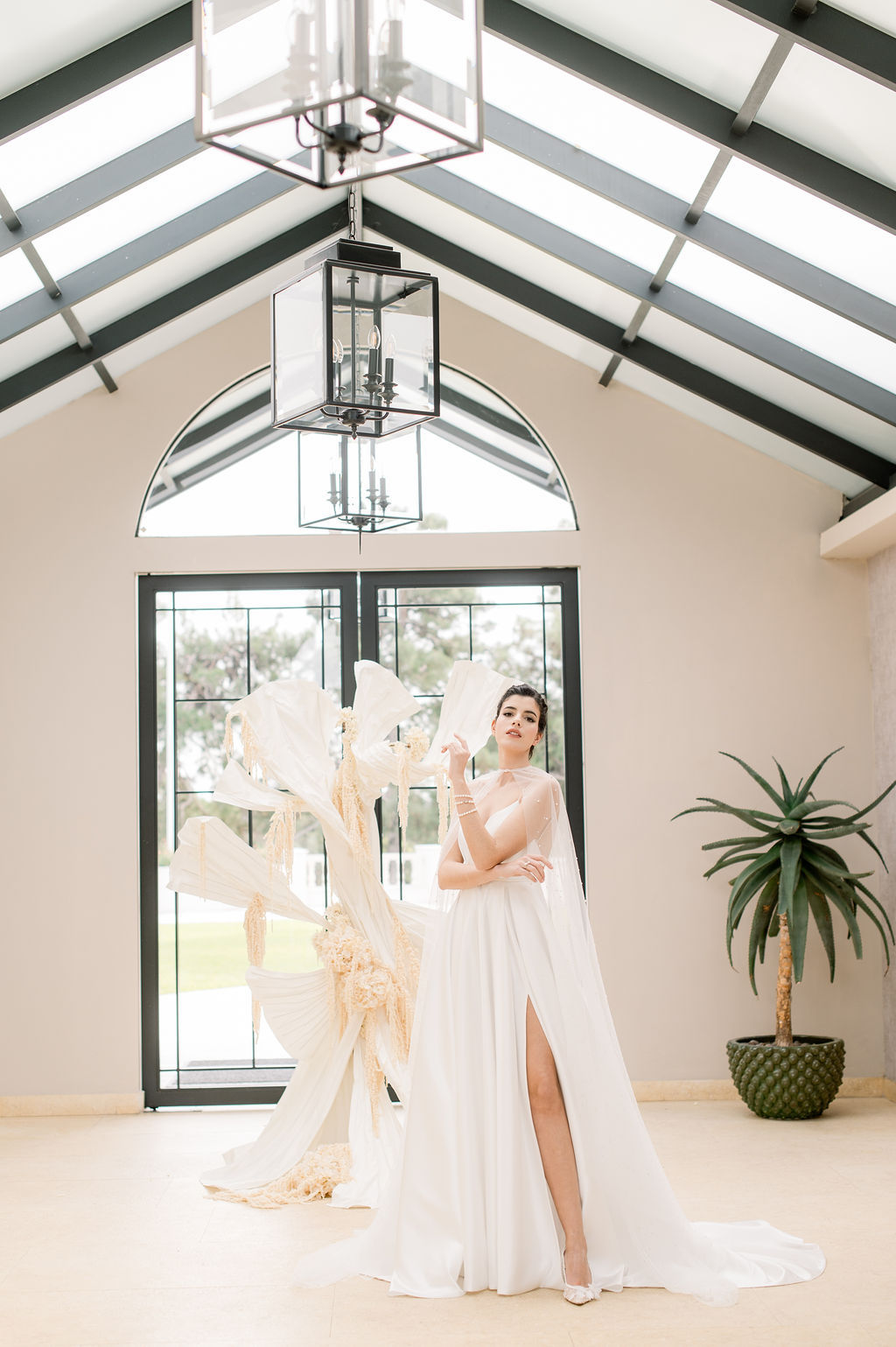 A woman in a white, sleeveless gown with a high slit stands indoors near large windows, holding a fan, with decorative white structures and a potted plant in the background.