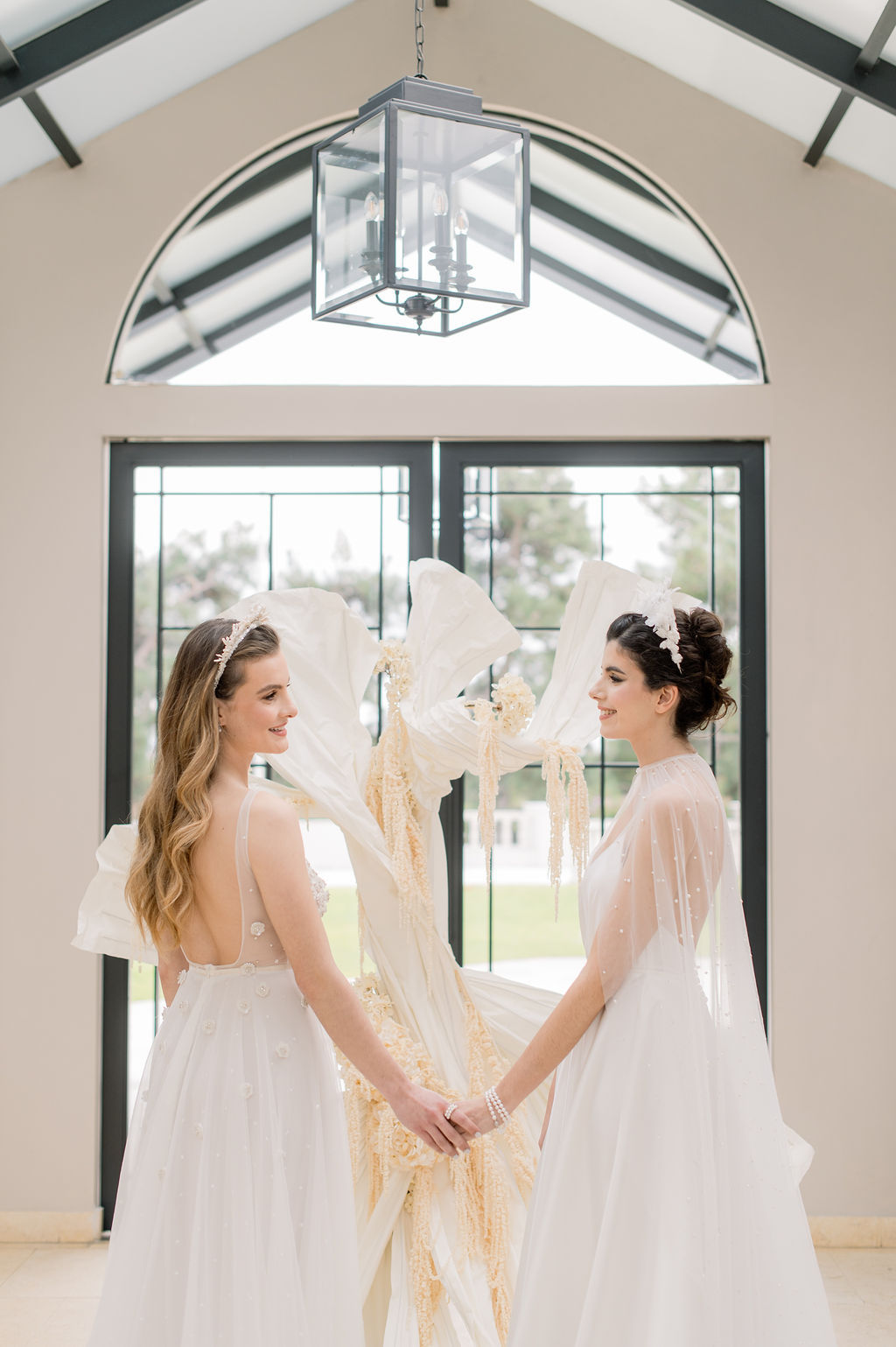 Two women in white wedding dresses stand facing each other, holding hands, in front of a large window with natural light streaming in.
