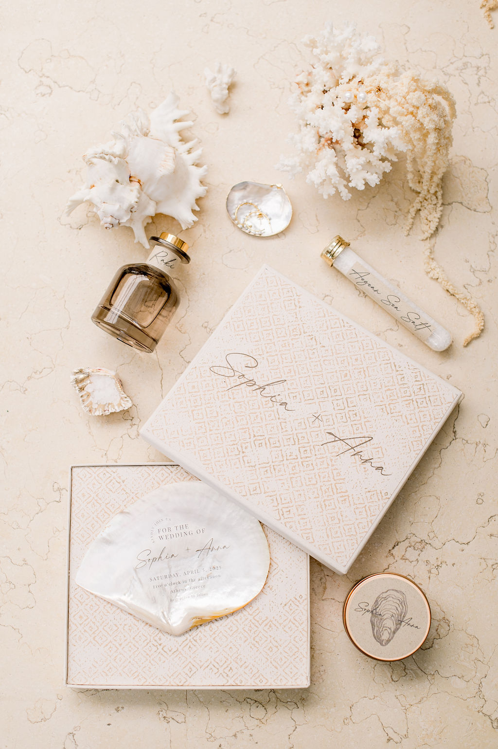 A flat lay of gift packaging with a box, shell, perfume bottle, test tube, and coral decorations arranged on a textured beige surface.