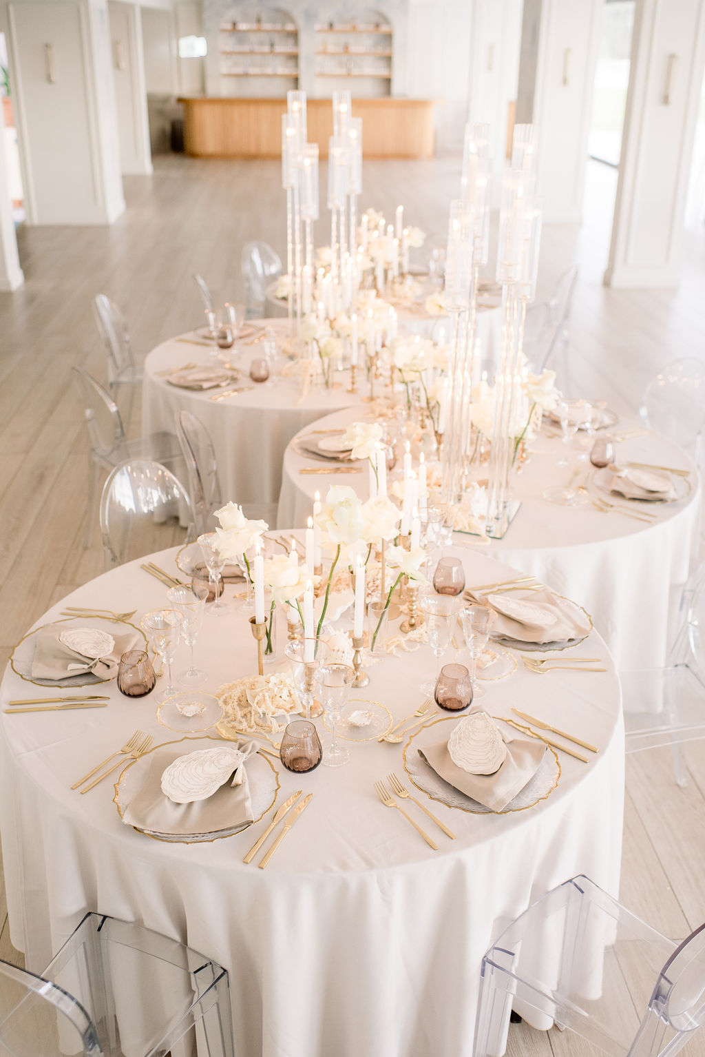 Elegant round tables set with white linens, gold cutlery, clear plates, and tall glass candle holders in a bright, modern event space with clear chairs.