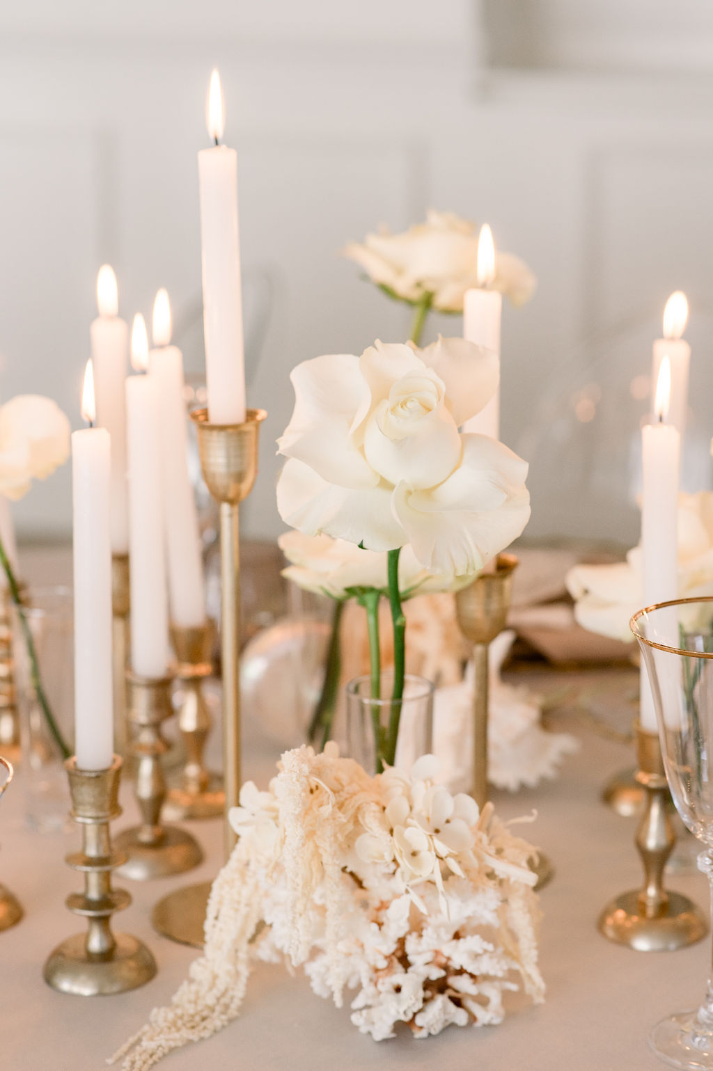 White roses in glass vases and clusters of white flowers are arranged with lit white candles in brass holders on a table.