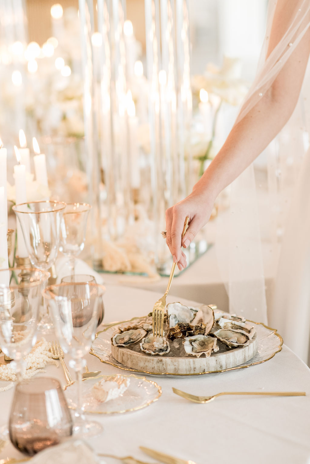 A person uses a fork to pick up an oyster from a platter on an elegant table set with candles, glassware, and gold utensils.