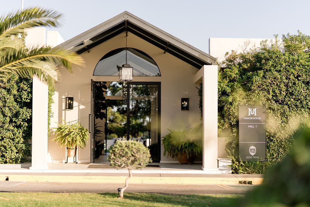 Modern building entrance with glass double doors, potted plants on either side, and greenery surrounding the exterior.