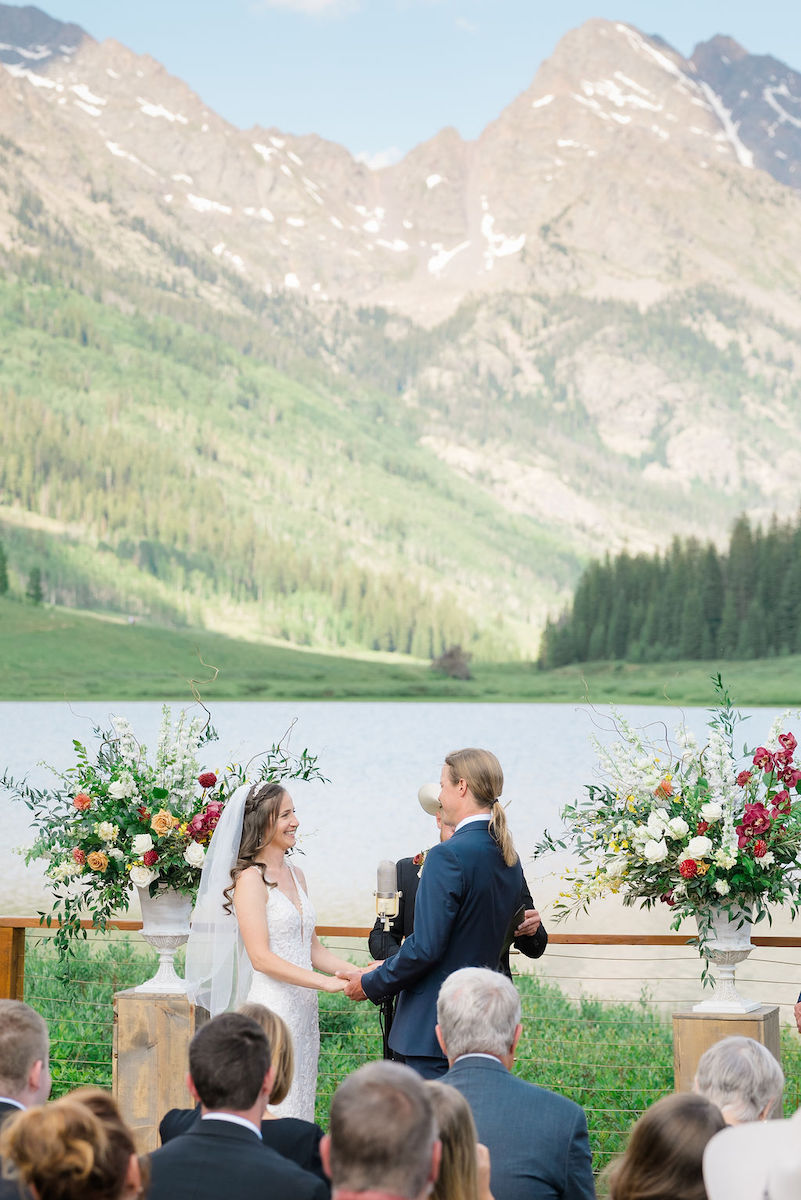 A wedding ceremony takes place outdoors in front of a scenic mountain landscape, with the bride and groom holding hands and guests seated in the foreground.