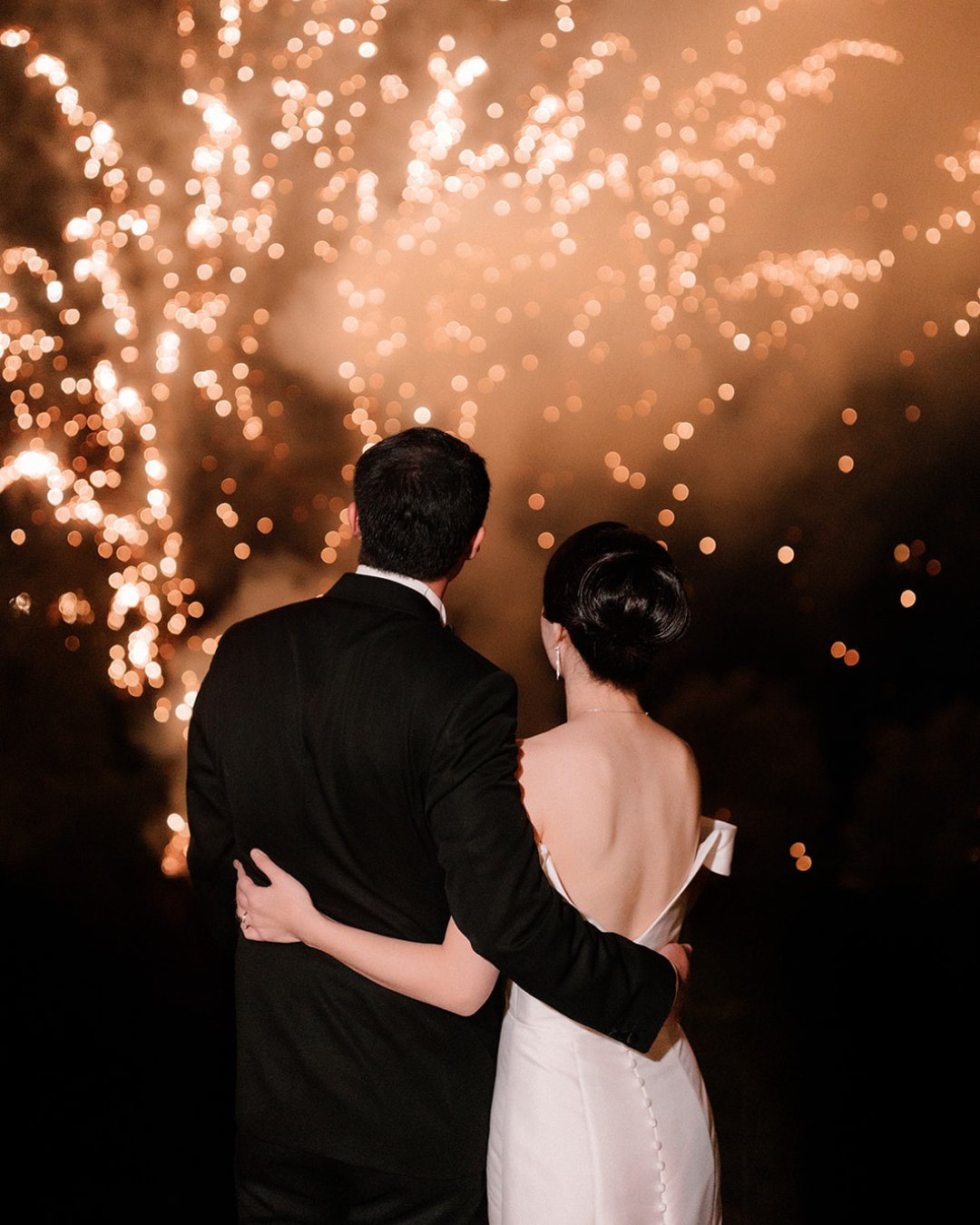 A couple in formal attire stands embracing, watching a fireworks display at night.