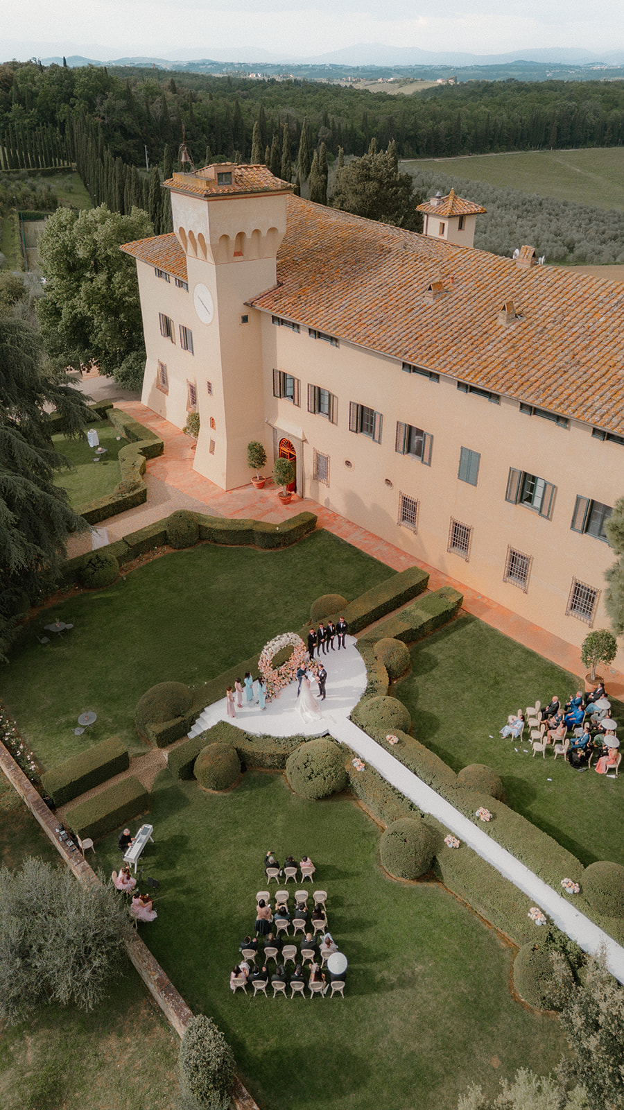 Aerial view of an outdoor wedding ceremony on the lawn of a large, historic villa with guests seated and trees surrounding the area.