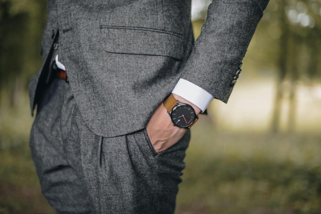 A person wearing a grey suit with their right hand in their pocket, showing a brown leather wristwatch.