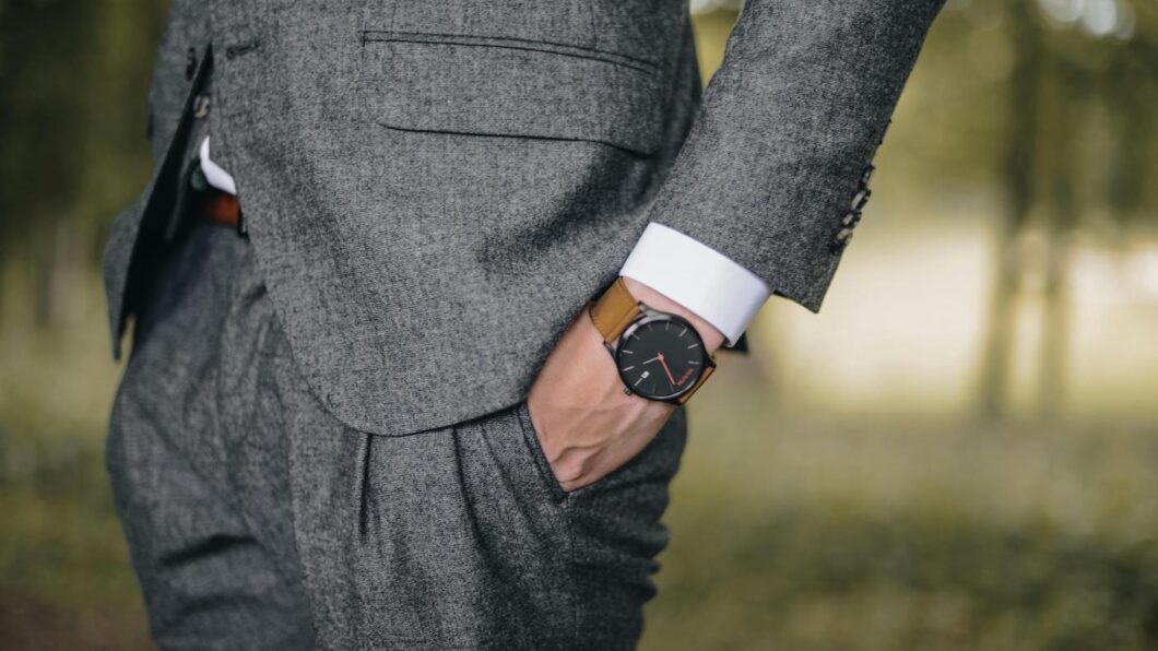A person wearing a grey suit with their right hand in their pocket, showing a brown leather wristwatch.