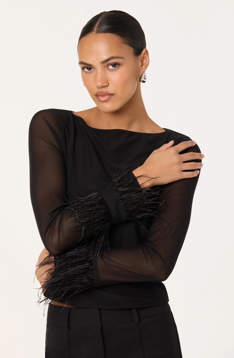 A woman wearing a sheer black long-sleeve top with feathered cuffs stands with one arm crossed over her chest against a light background.