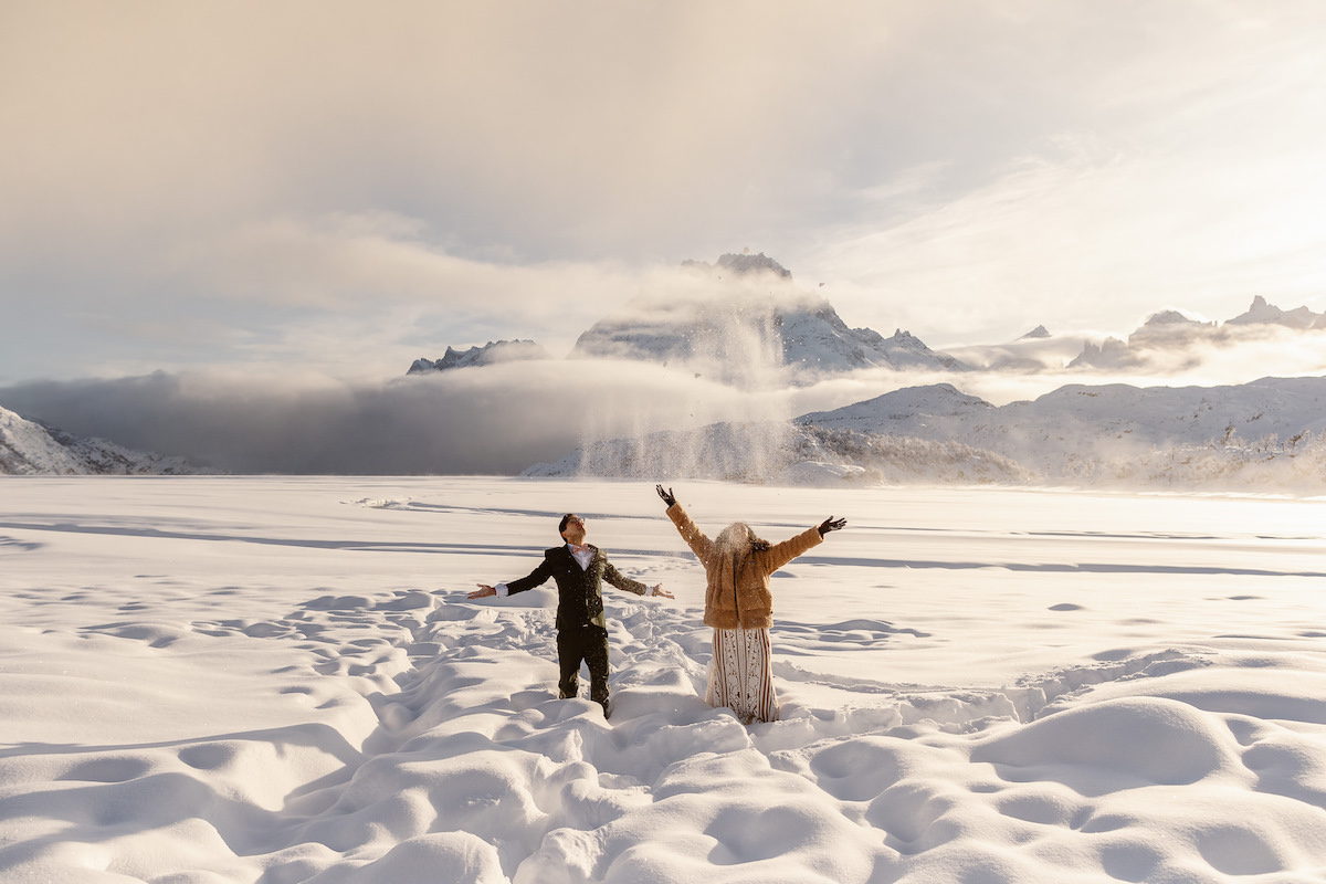 Two people stand in deep snow with arms outstretched, surrounded by a snowy landscape and distant mountains under a cloudy sky.