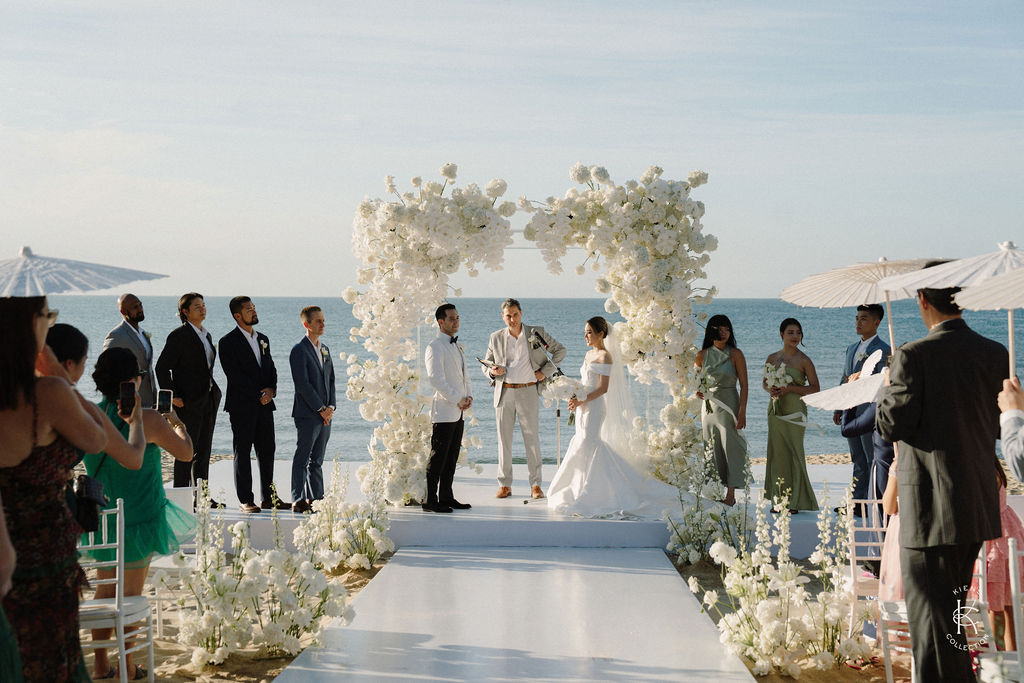 A wedding ceremony takes place on a beach, with the couple standing under a white floral arch, surrounded by bridesmaids, groomsmen, and guests.