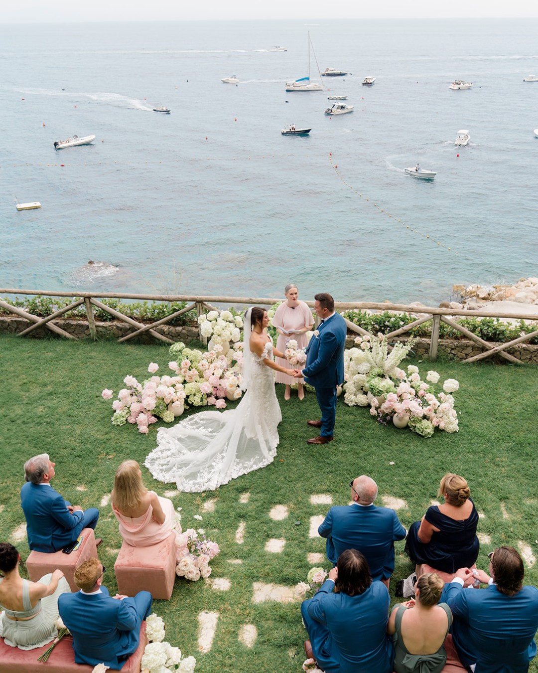 A bride and groom stand with an officiant on a grassy area decorated with flowers, overlooking the ocean, as a small group of guests watch the ceremony.