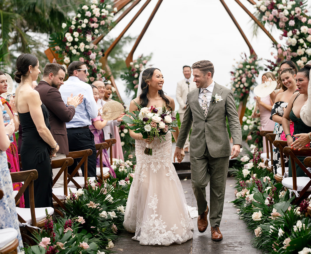 A bride and groom walk down the aisle together, smiling, surrounded by guests clapping and floral decorations at an outdoor wedding ceremony.