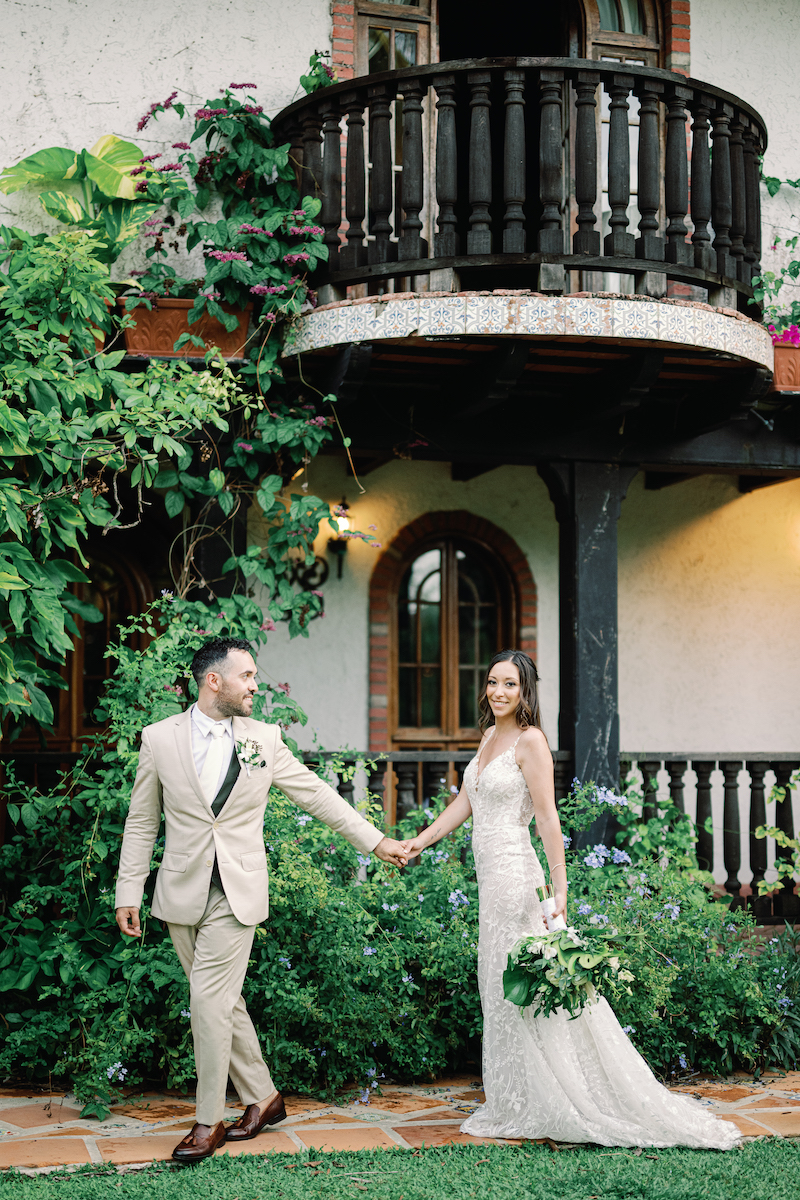 A bride and groom hold hands outside a rustic building with a balcony, surrounded by lush greenery and flowers.