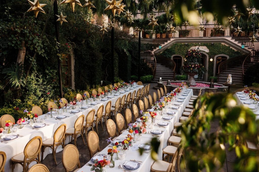 Long banquet tables with white tablecloths and gold chairs are set for an outdoor event, surrounded by greenery and star-shaped hanging decorations.