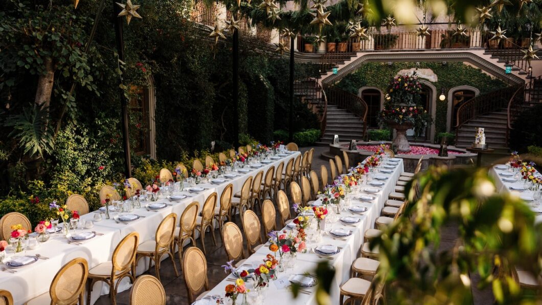Long banquet tables with white tablecloths and gold chairs are set for an outdoor event, surrounded by greenery and star-shaped hanging decorations.