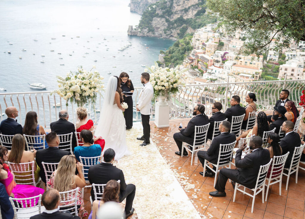 A bride and groom stand before an officiant at an outdoor wedding ceremony overlooking the sea, with guests seated in rows on a terrace.