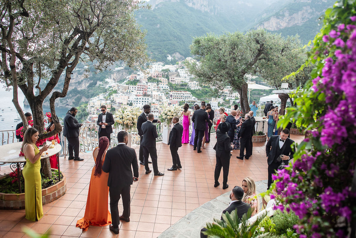 People dressed formally gather on a terrace with trees, flowers, and a scenic hillside town in the background.